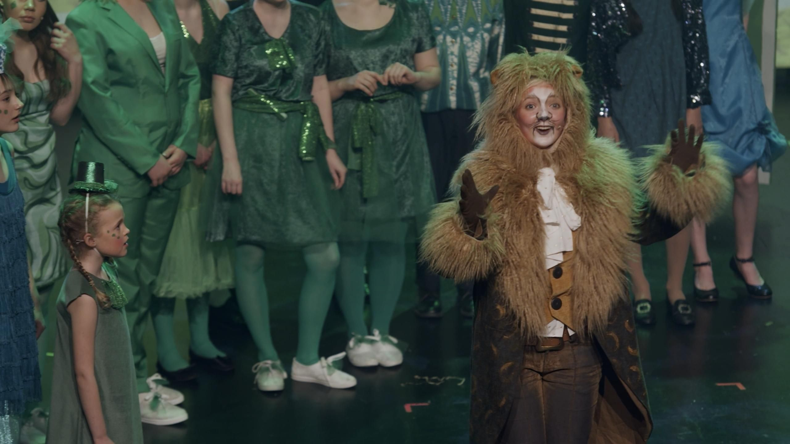 Den feige løven synger på scenen med folk kledd i grønt