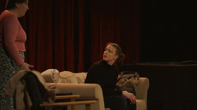Stående person prater med kvinne som sitter i sofa. Bilde fra forestilling