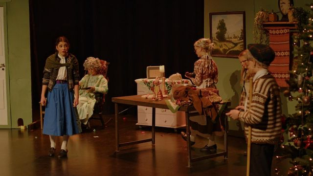 Barn i gammeldagse klær står på scene dekorert som gammel stue