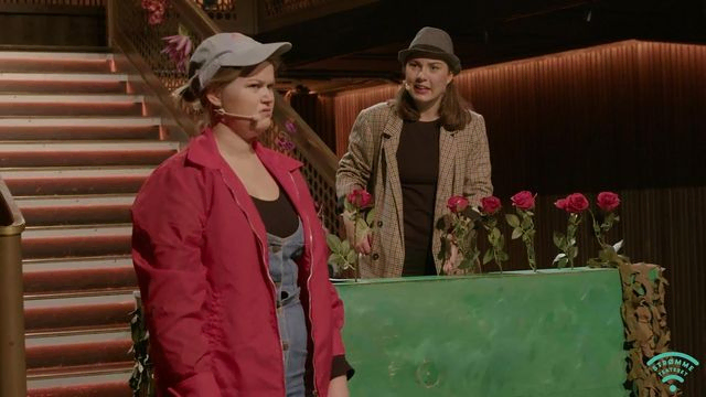 Person i rød jakke skjærer grimase, med detektiv Dora i bakgrunn med blomsterbedd av roser