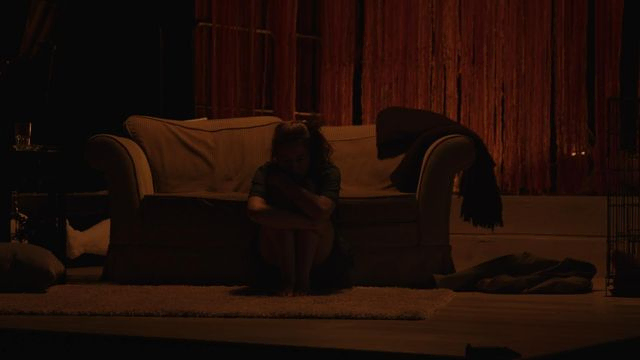 Person sitter på gulv foran sofa i mørket med hodet i fanget. Bilde fra forestilling